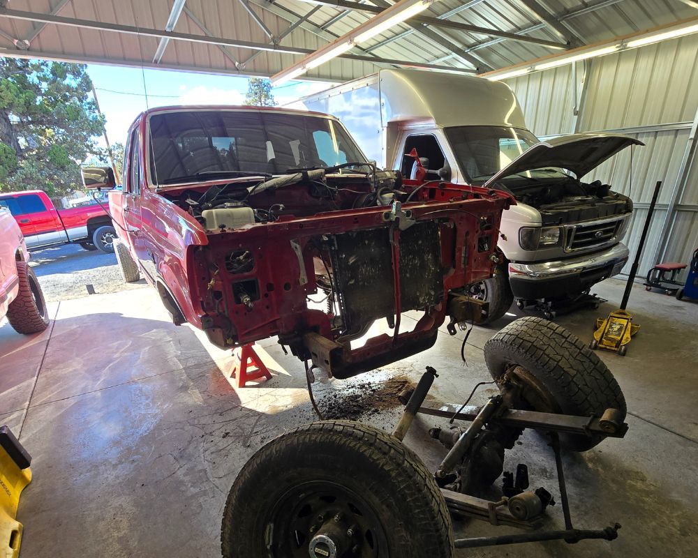 Red pickup truck body elevated on jack stands with front axle separated in a garage with a white truck nearby and various tools around.