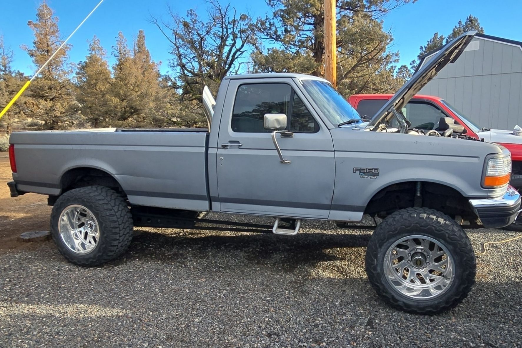 Light gray lifted Ford F-350 XL pickup truck with large off-road tires and open hood parked on gravel in front of trees and a building.